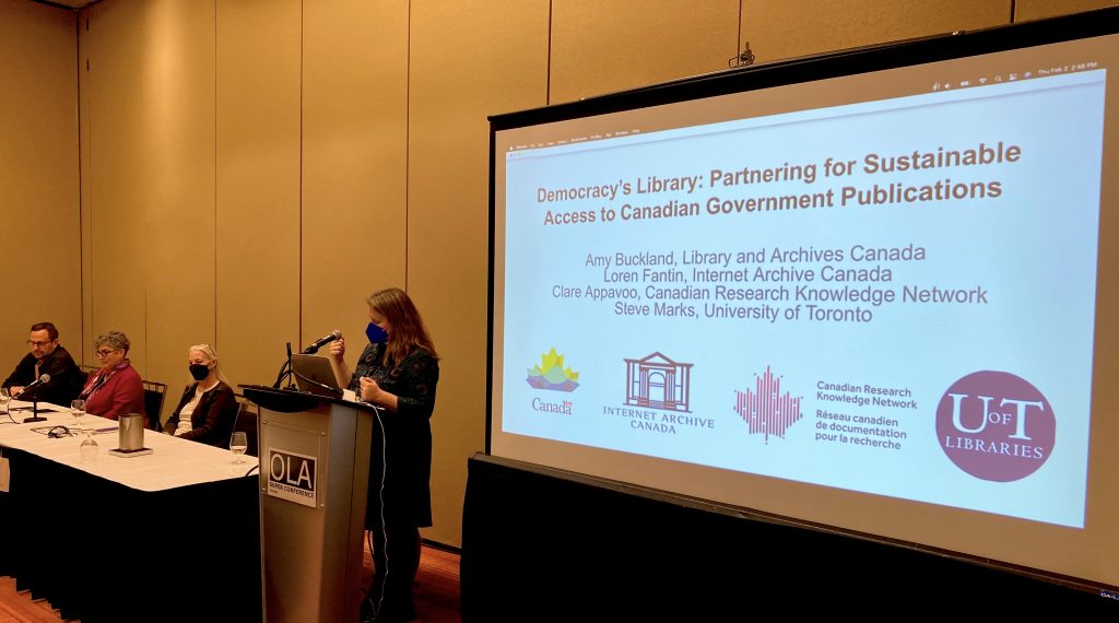 Conference presentation. 3 people sitting at a panel table and one person standing at a podium, speaking. The podium is marked OLA and the screen is showing a slide titled: " Democracy's Library: Partnering for Sustainable Access to Canadian Government Publications".